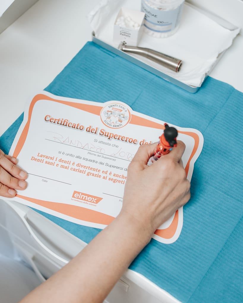 Una mano compila il Certificato del Supereroe dei Denti sul tavolo del riunito, TCL Dental
