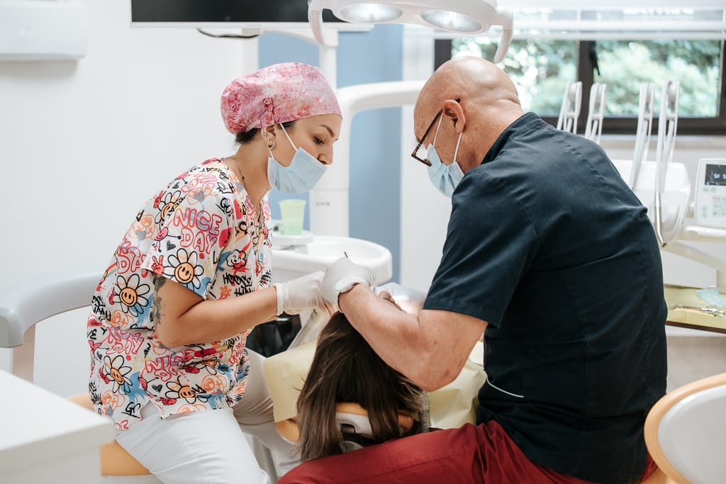Due operatori con mascherina e cuffia al lavoro su un paziente sotto la lampada del riunito, TCL Dental Barcellona Pozzo di Gotto