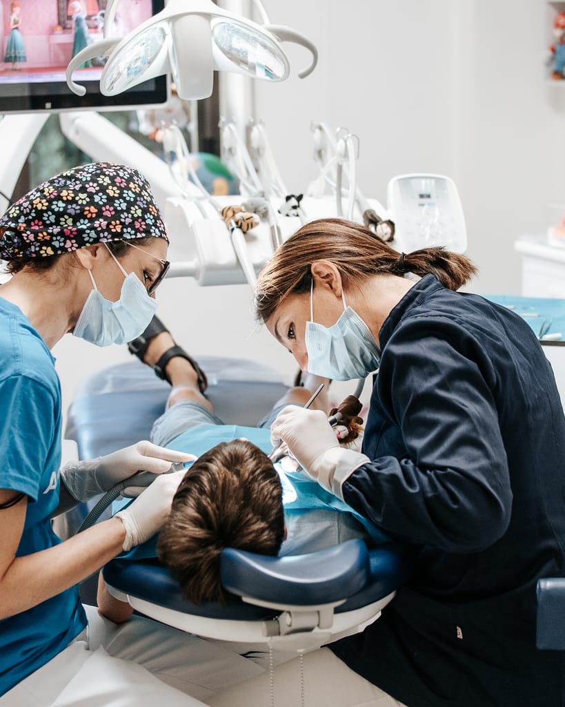 Due operatrici in divisa azzurra chinate su un piccolo paziente, dettaglio delle mani con gli strumenti